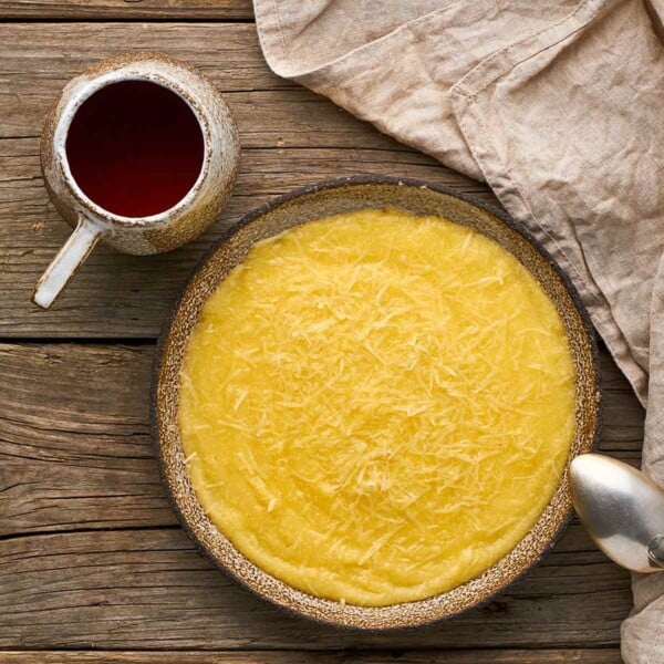 A bowl filled with basic polenta next to a mug of tea, a cloth, and a spoon resting on the side of the bowl.