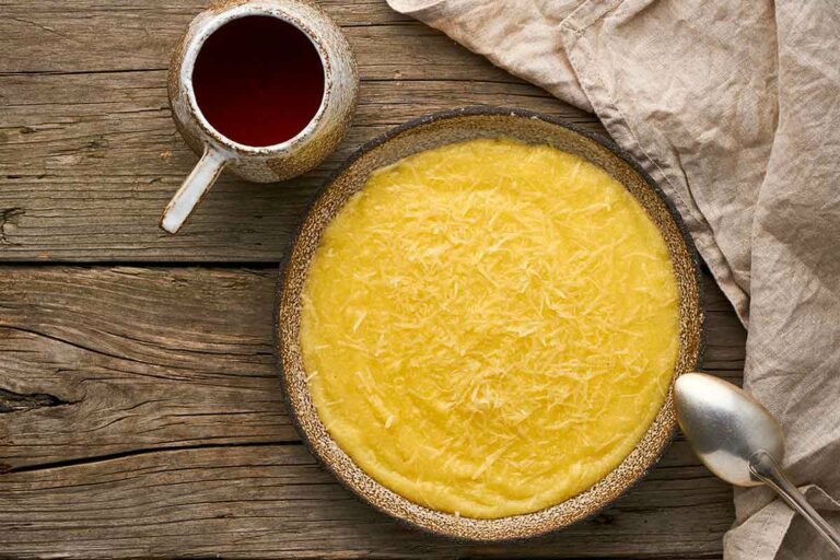 A bowl filled with basic polenta next to a mug of tea, a cloth, and a spoon resting on the side of the bowl.