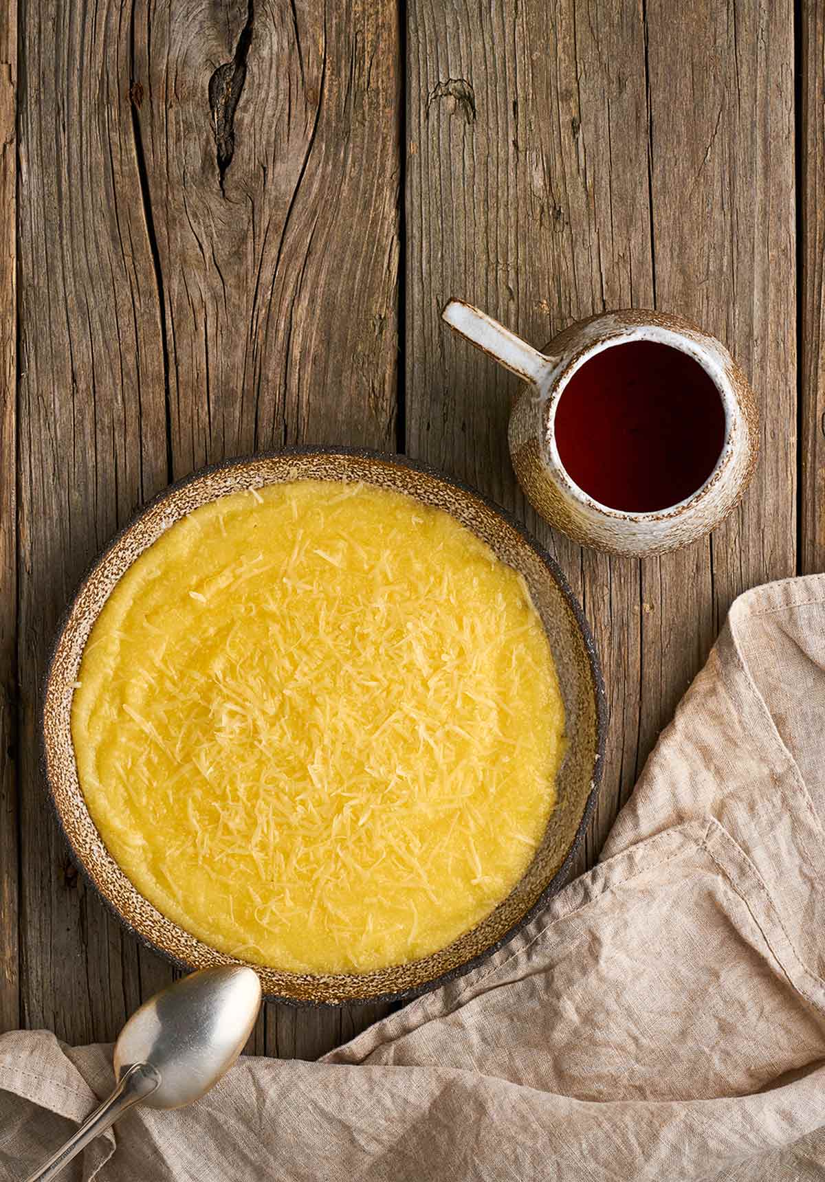 A bowl filled with basic polenta next to a mug of tea, a cloth, and a spoon resting on the side of the bowl.