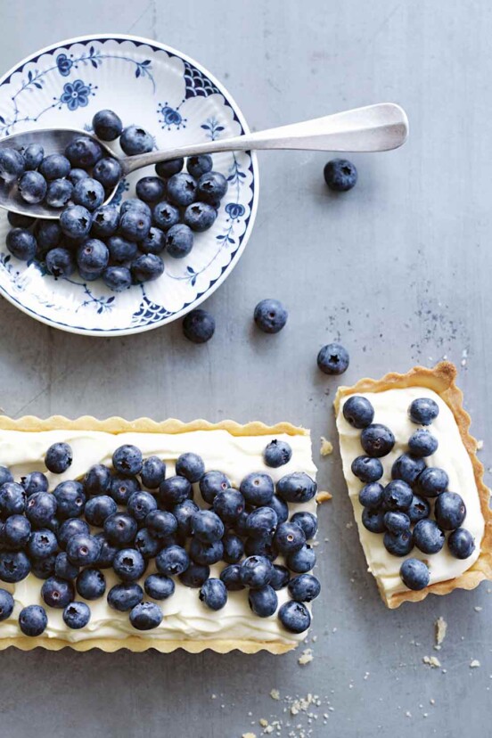 A rectangular blueberry mascarpone tart with one slice cut from the end and a bowl of blueberries on the side.