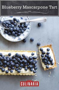 A rectangular blueberry mascarpone tart with one slice cut from the end and a bowl of blueberries on the side.