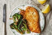 A piece of bread crumb coated chicken on a patterned plate with broccoli and a fork and two lemon halves on the side.