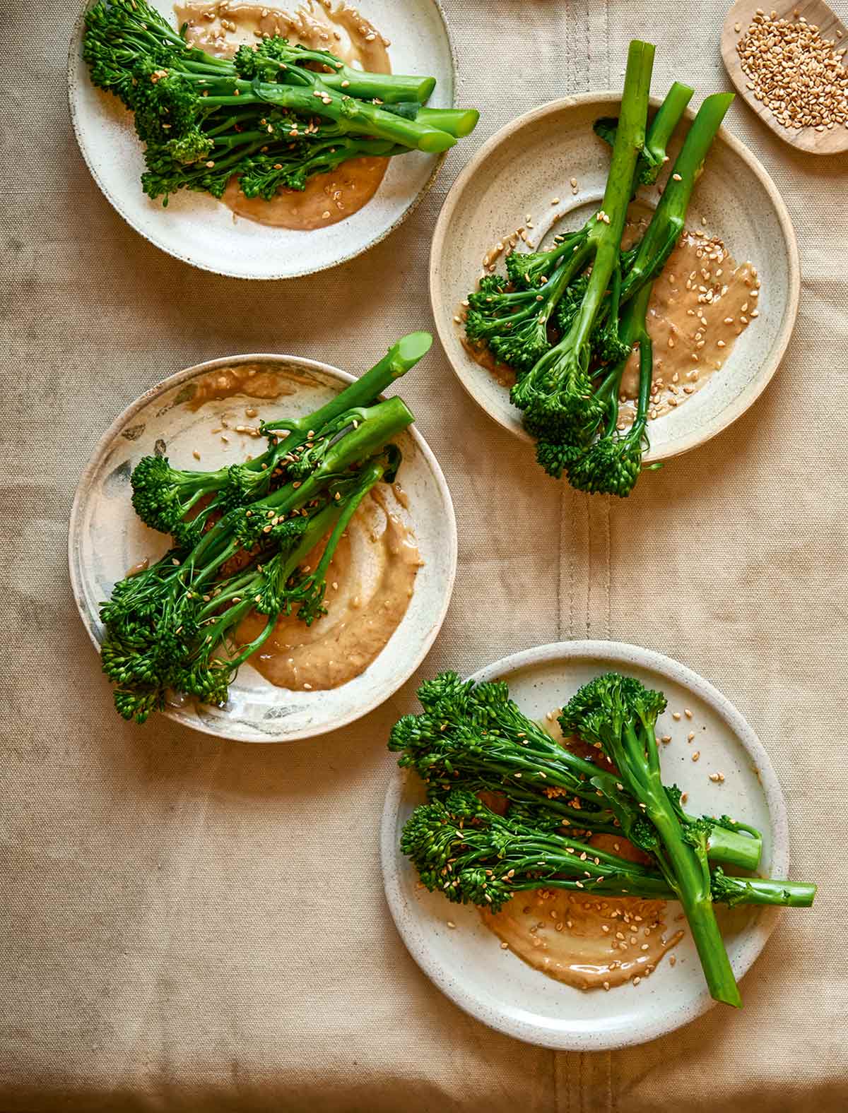 Four plates topped with broccoli with peanut sauce and a small dish of sesame seeds on the side.