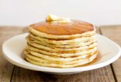 A stack of buttermilk pancakes on a white plate with a pat of butter on top and syrup running down the sides.