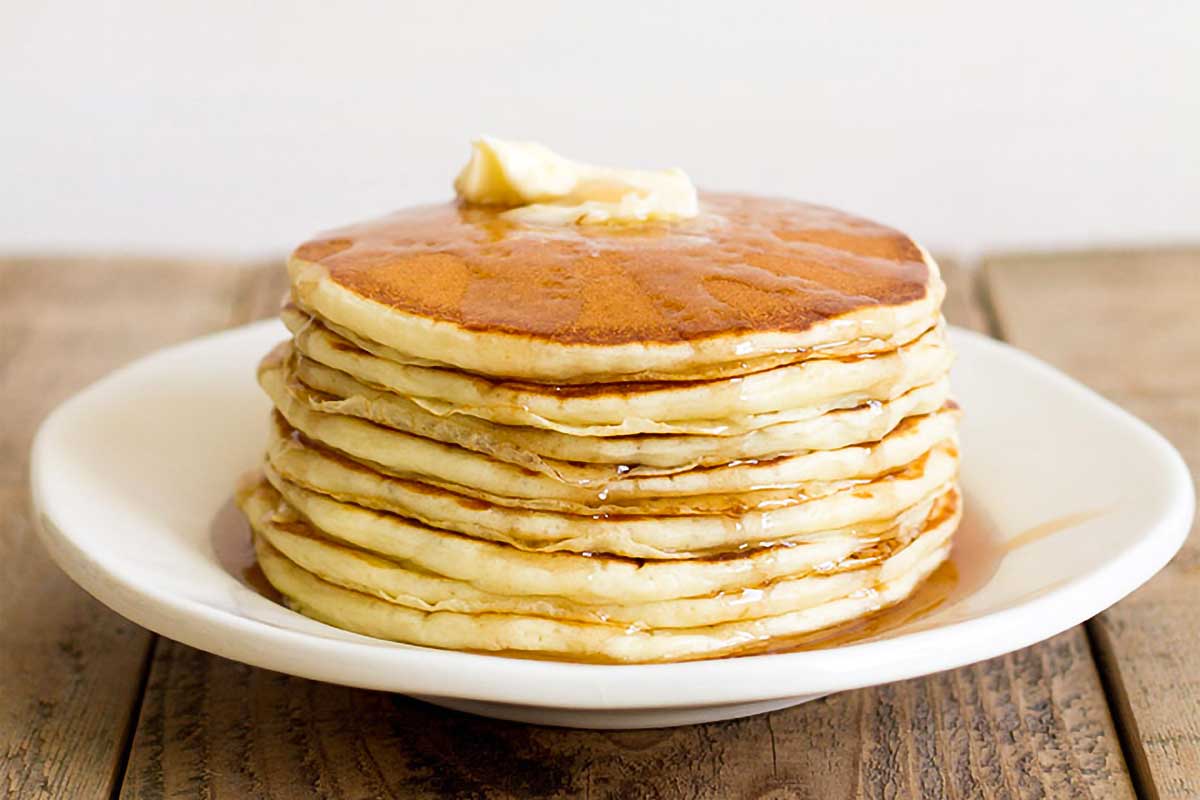 A stack of buttermilk pancakes on a white plate with a pat of butter on top and syrup running down the sides.