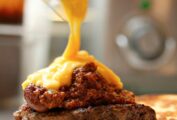Cheddar cheese sauce being ladled over a chili-topped burger.