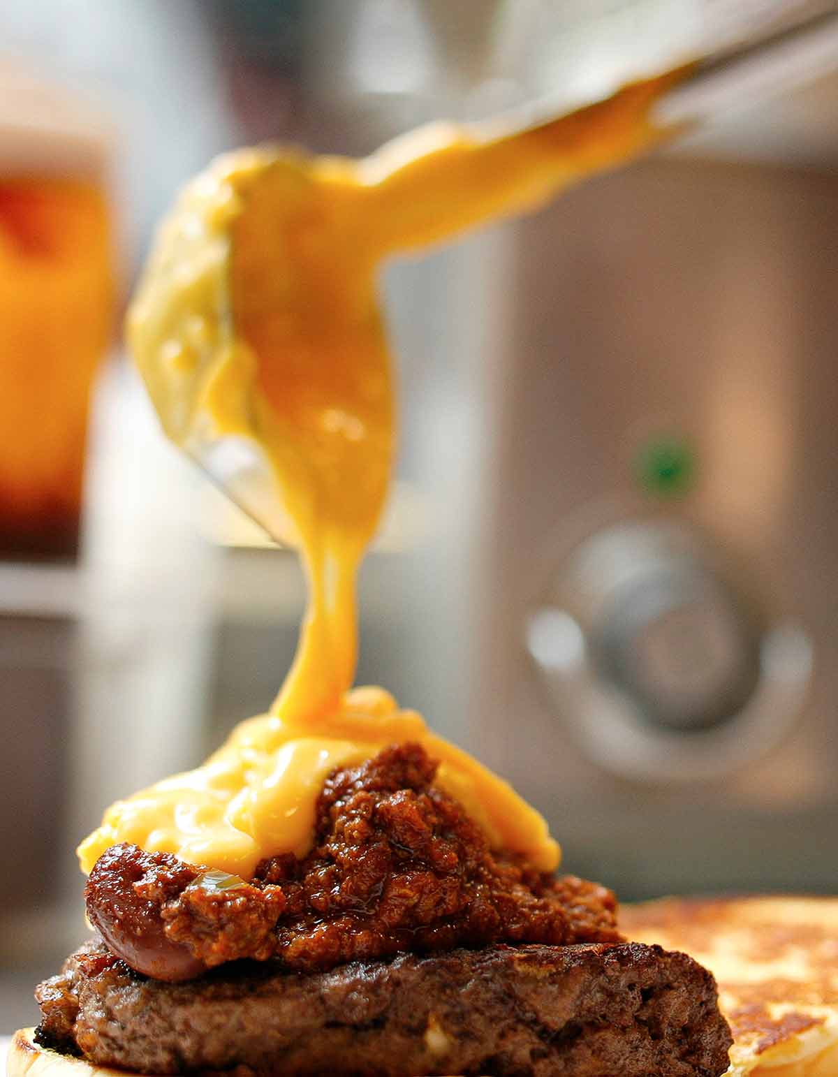 Cheddar cheese sauce being ladled over a chili-topped burger.