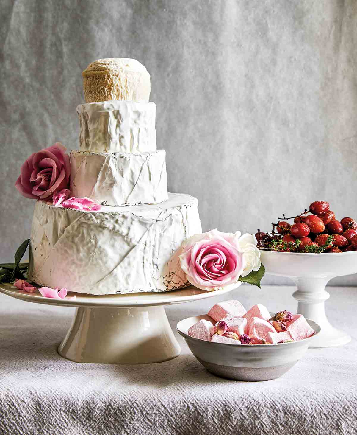 A cheese "wedding" cake made with four rounds of cheese, fresh roses, and bowls of roasted grapes and marshmallows on the side.