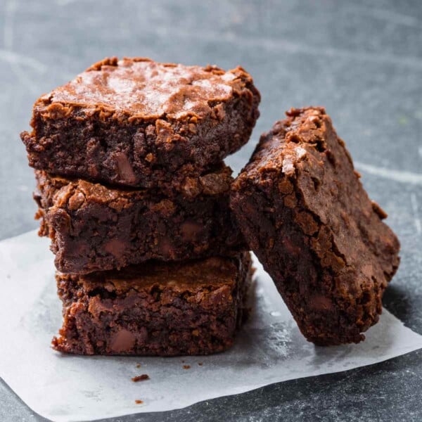 Three chewy chocolate brownies stacked on top of each other with another leaning on the stack on a piece of parchment.