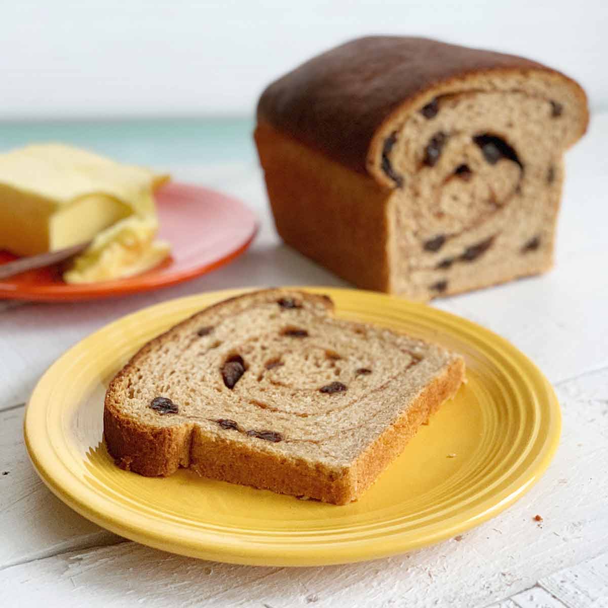A cut loaf of cinnamon raisin swirl bread and a slice of it on a yellow plate with butter in the background