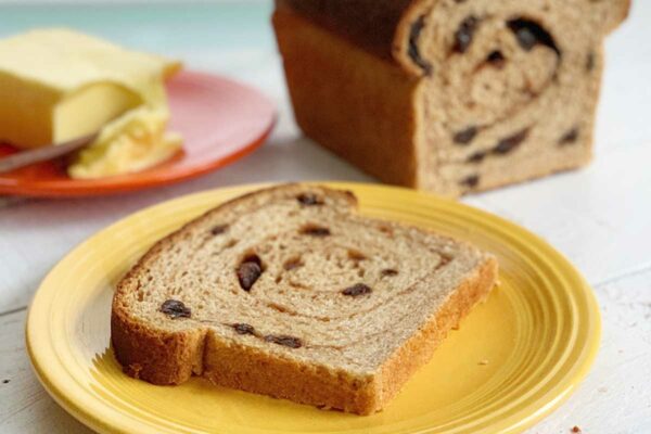 A cut loaf of cinnamon raisin swirl bread and a slice of it on a yellow plate with butter in the background