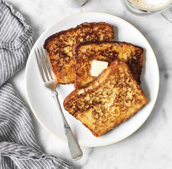 Three slices of classic French toast on a white plate with a pat of butter on top and a fork on the side.