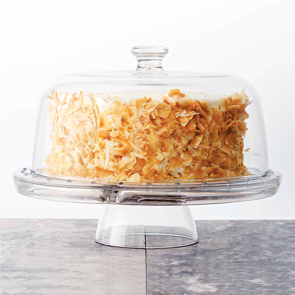A whole coconut carrot cake, covered in toasted coconut on a glass cake stand, covered with a glass dome.