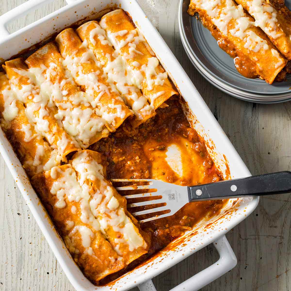 A rectangular dish of easy chicken enchiladas with a spatula resting in the dish and a couple enchiladas on a grey plate beside it.