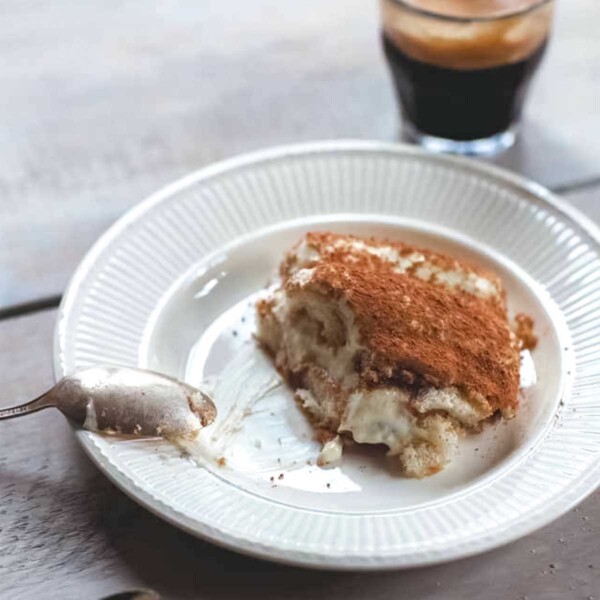A serving of easy tiramisu on a white plate with a spoon resting on the side and a shot of espresso in the background.