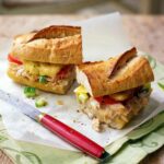A halved fancy tuna melt with tomato and cucumber on a baguette on a piece of parchment.