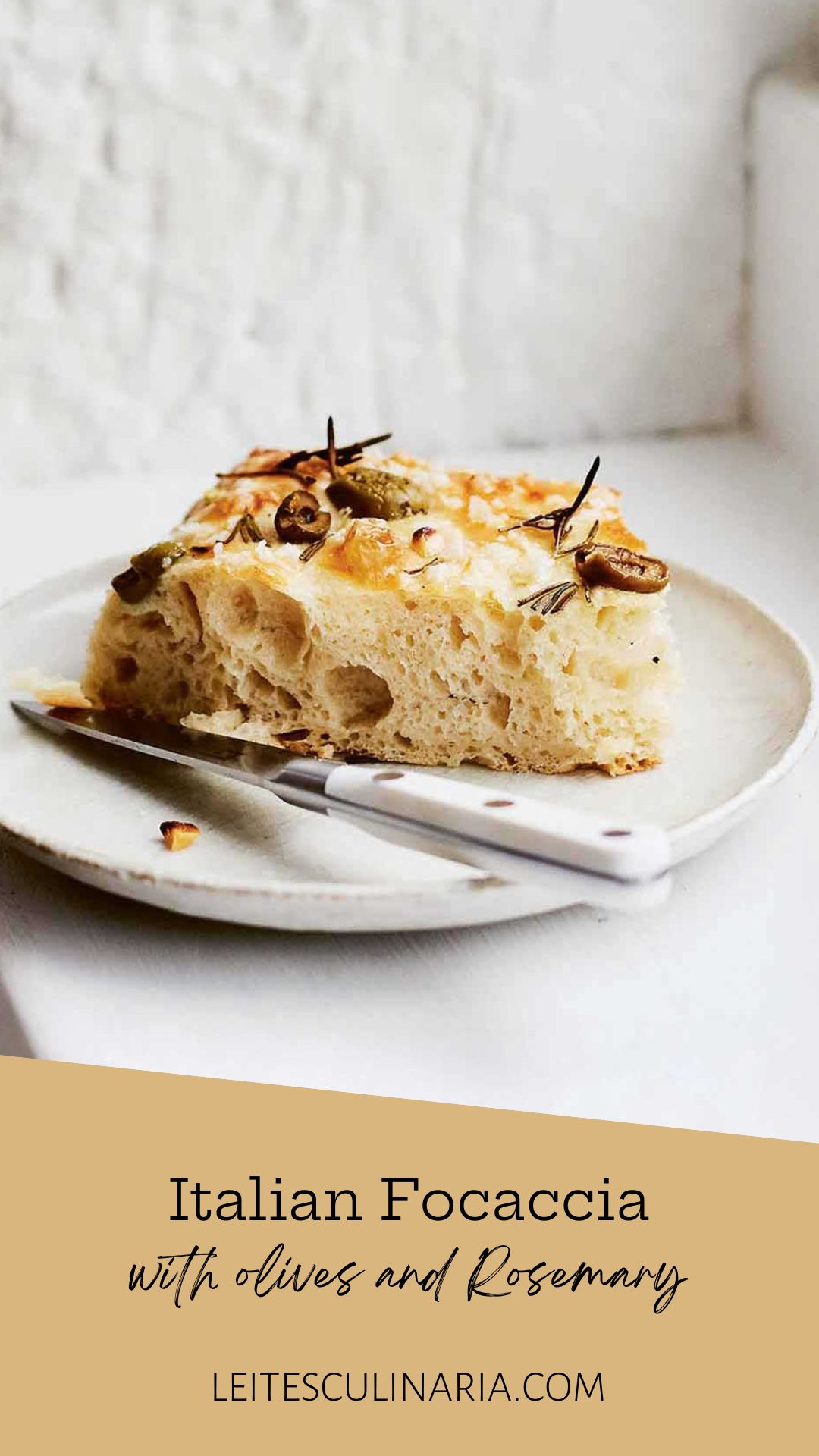 A slice of focaccia with olives and rosemary on a white plate with a knife resting beside it.