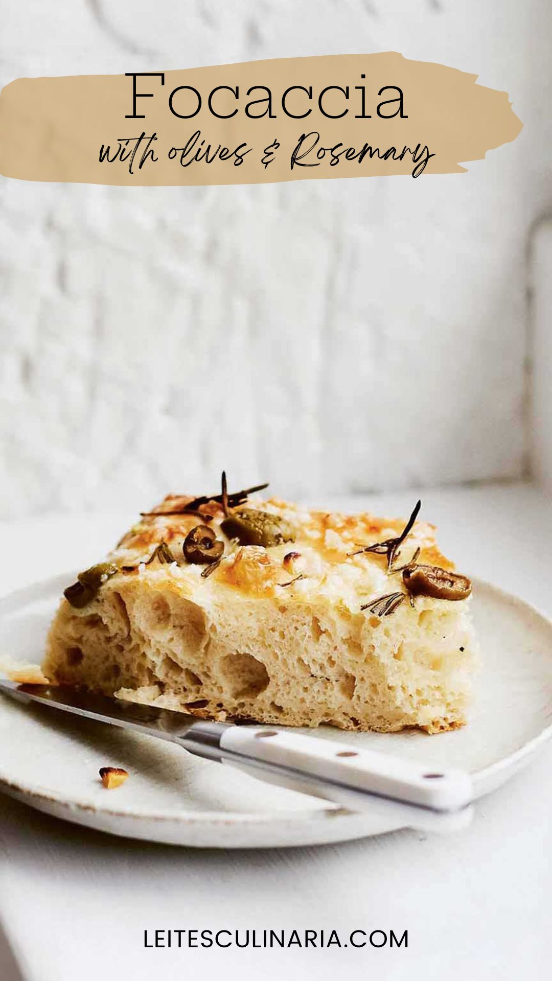 A slice of focaccia with olives and rosemary on a white plate with a knife resting beside it.