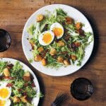 Two white plates of frisée with bacon and egg, croutons, and a dressing, glasses and fork and knife nearby