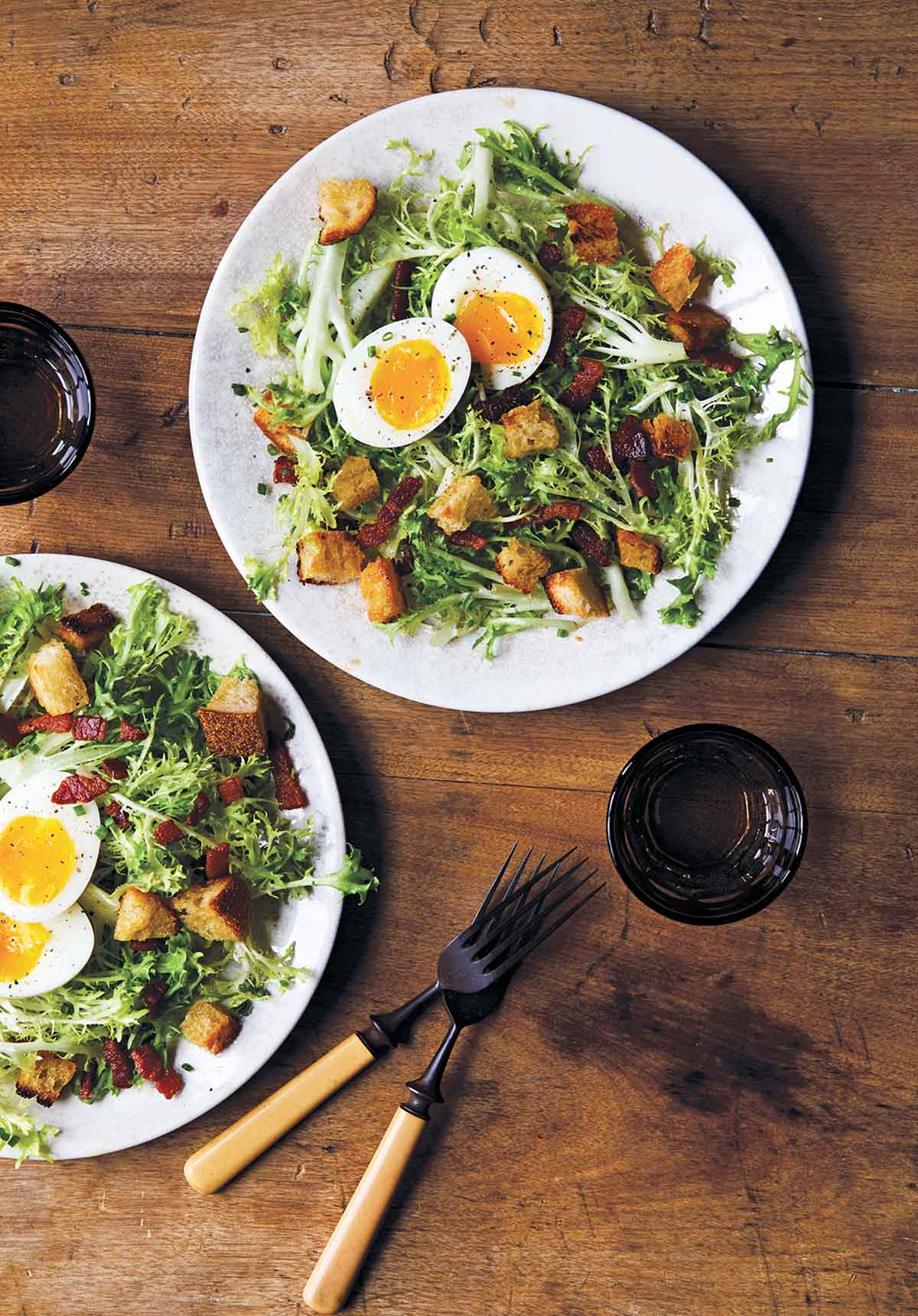 Two white plates of frisée with bacon and egg, croutons, and a dressing, glasses and fork and knife nearby