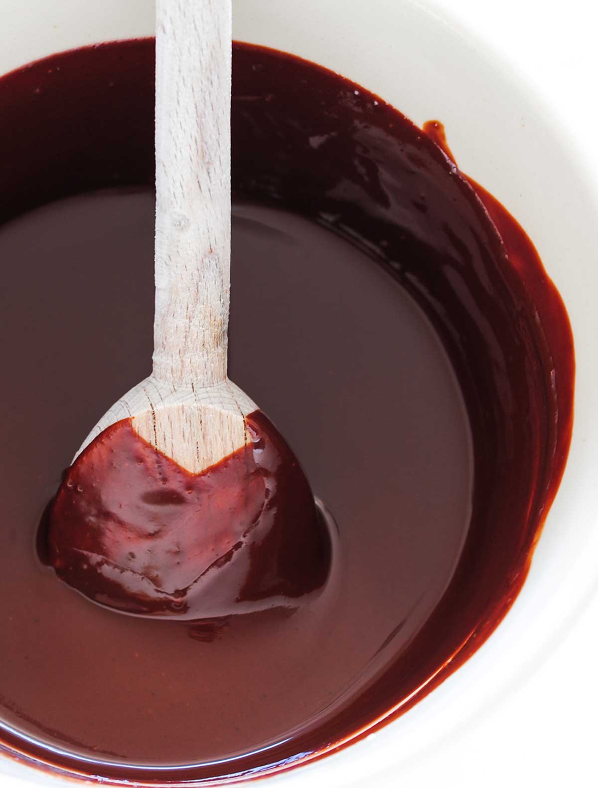 A white bowl filled with chocolate ganache and a wooden spoon resting inside.