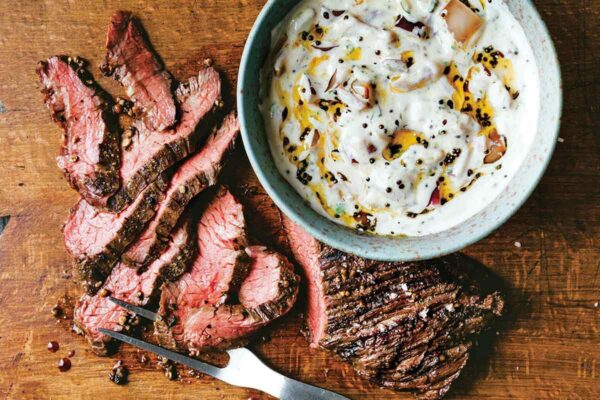 A sliced grilled hanger steak with raita in a bowl next to it on a wooden cutting board.