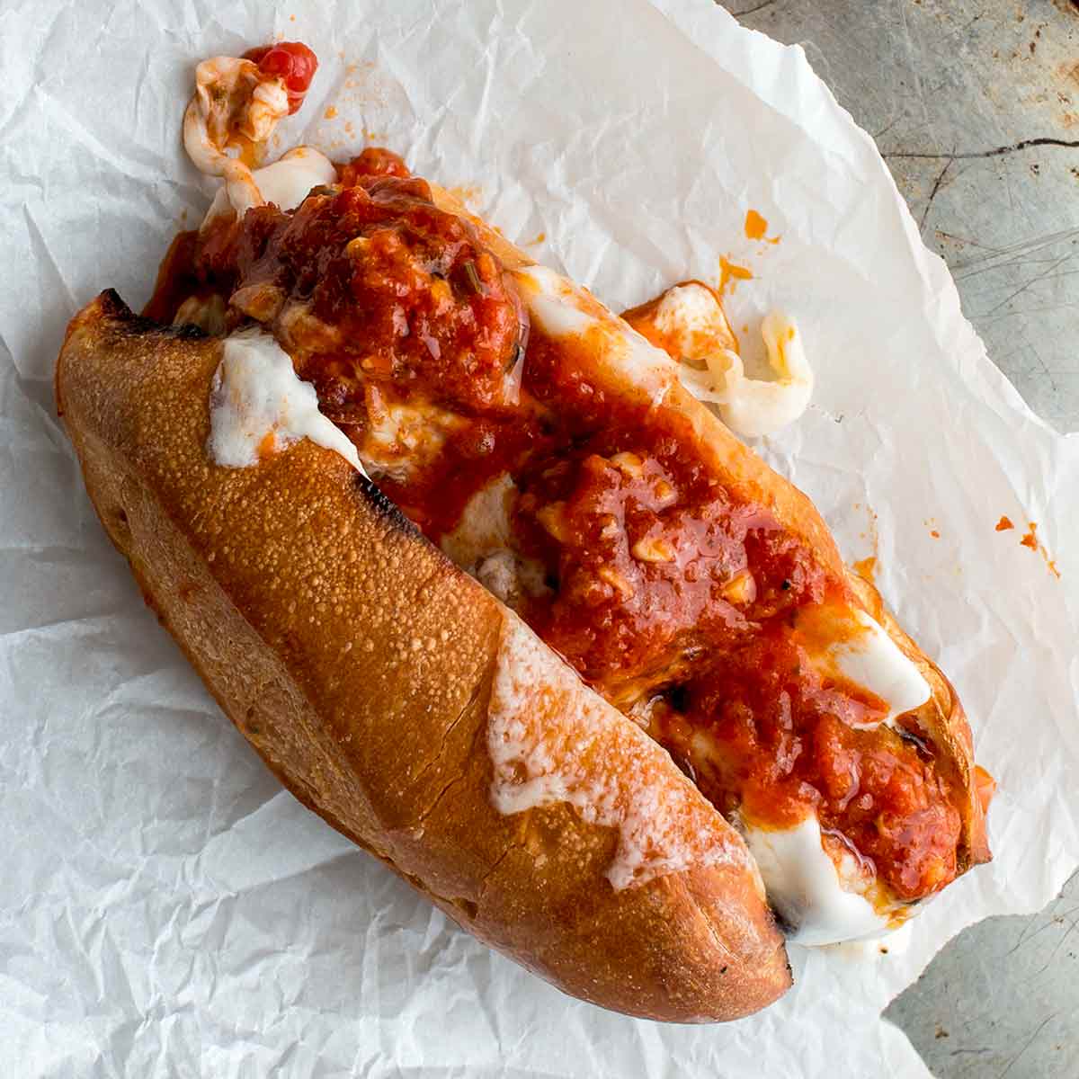 A harissa meatballs sandwich, made with three meatballs stuffed in a sub roll along with cheese and topped with tomato sauce on a sheet of parchment paper.