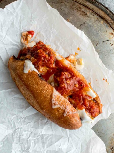 A harissa meatballs sandwich, made with three meatballs stuffed in a sub roll along with cheese and topped with tomato sauce on a sheet of parchment paper.