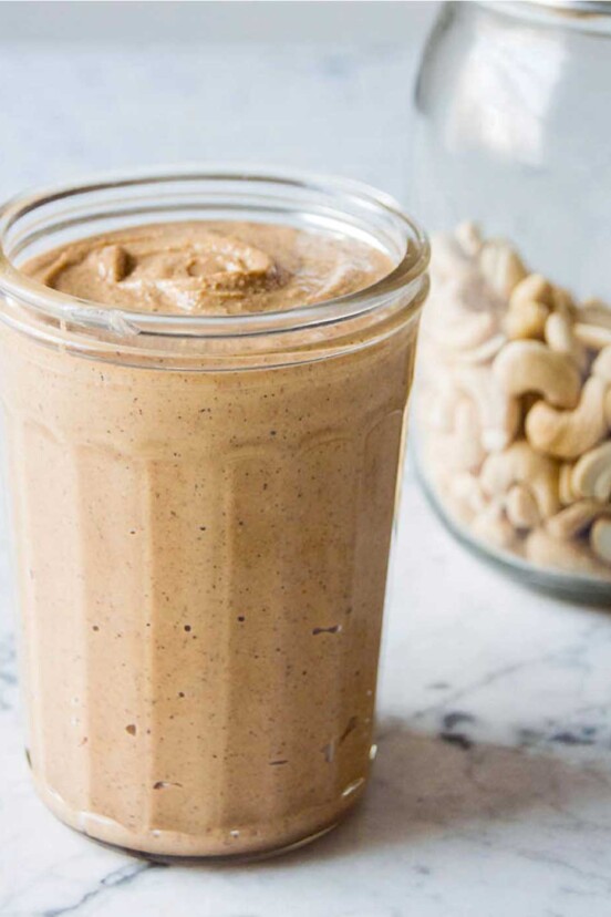 A glass jar filled with homemade cashew butter with a jar of cashews in the background.