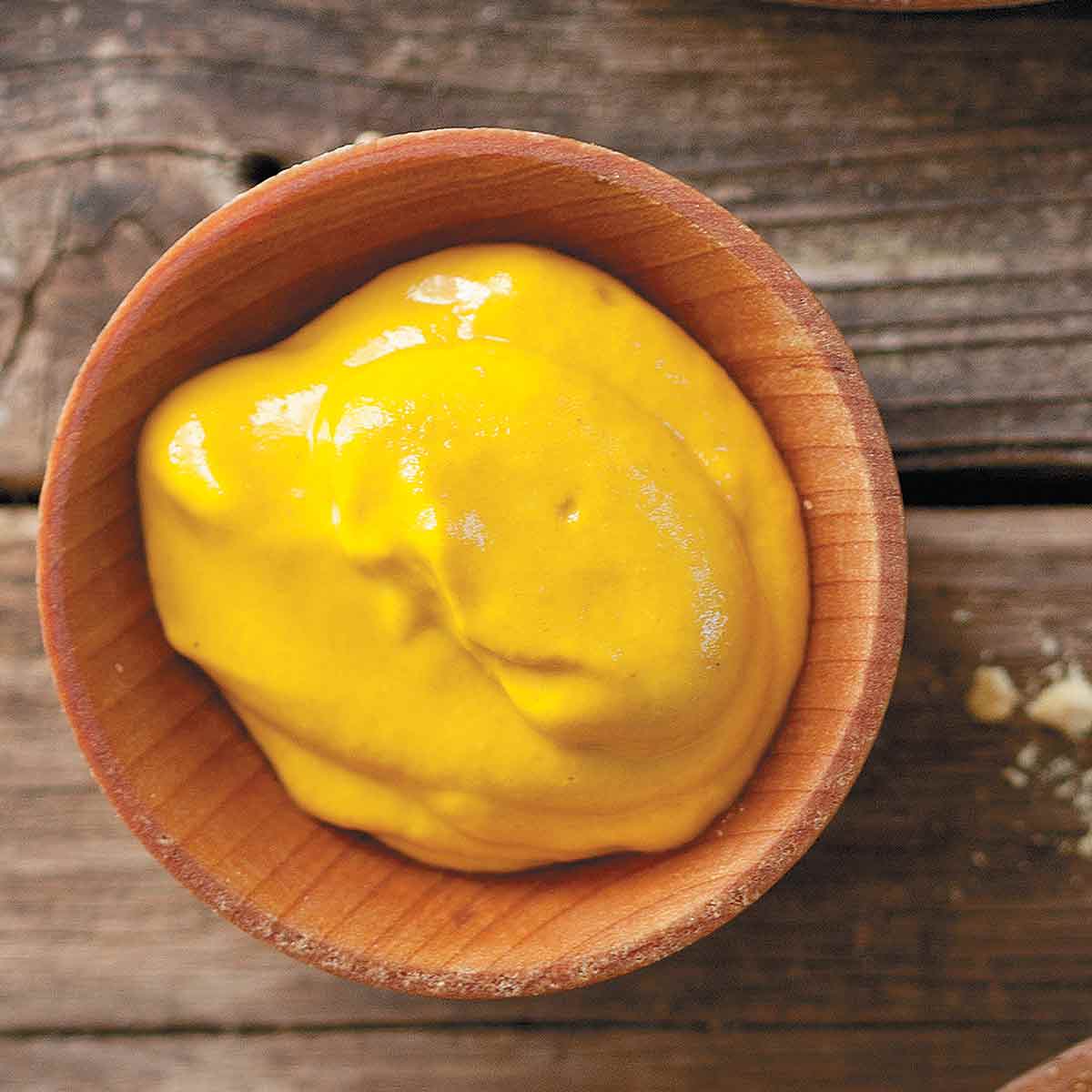 A bowl of homemade yellow mustard next to a wooden spoon filled with mustard seeds.