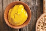A bowl of homemade yellow mustard next to a wooden spoon filled with mustard seeds.