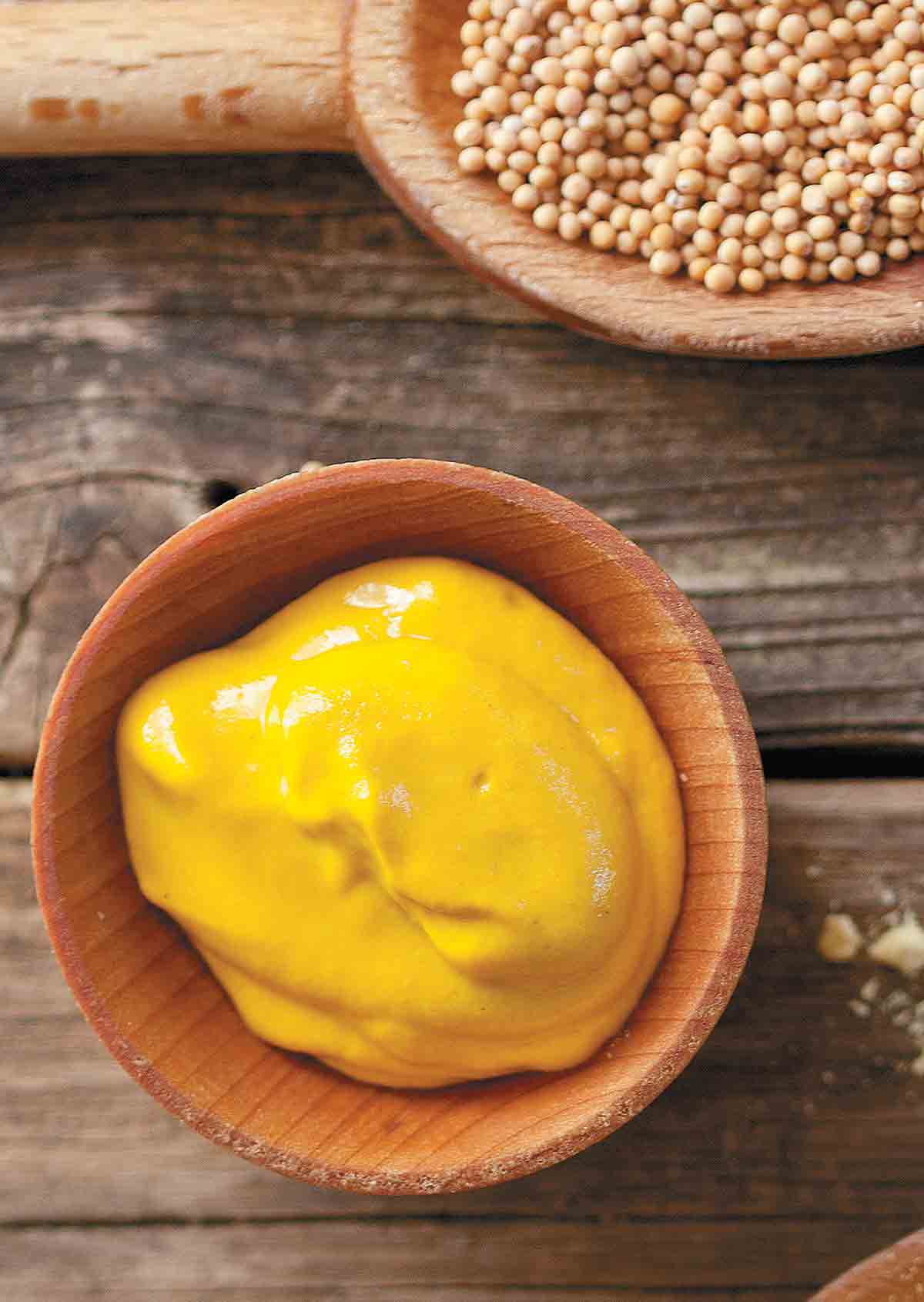 A bowl of homemade yellow mustard next to a wooden spoon filled with mustard seeds.