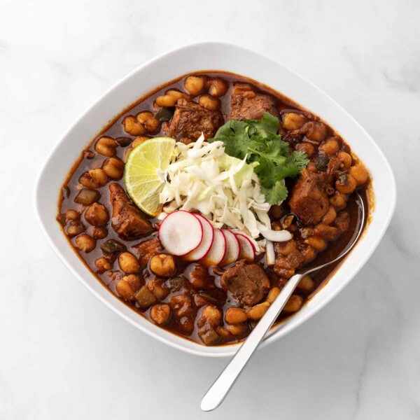 A square white bowl filled with Instant Pot pork pozole, garnished with radish, shredded cheese, lime, and cilantro, with a spoon resting in it.