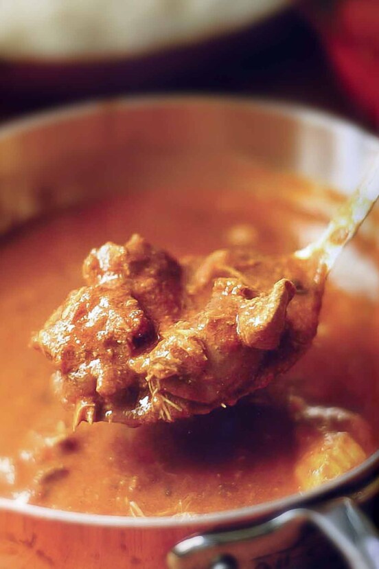 A spoon of a red chicken curry held above copper pot filled with the Indian stew