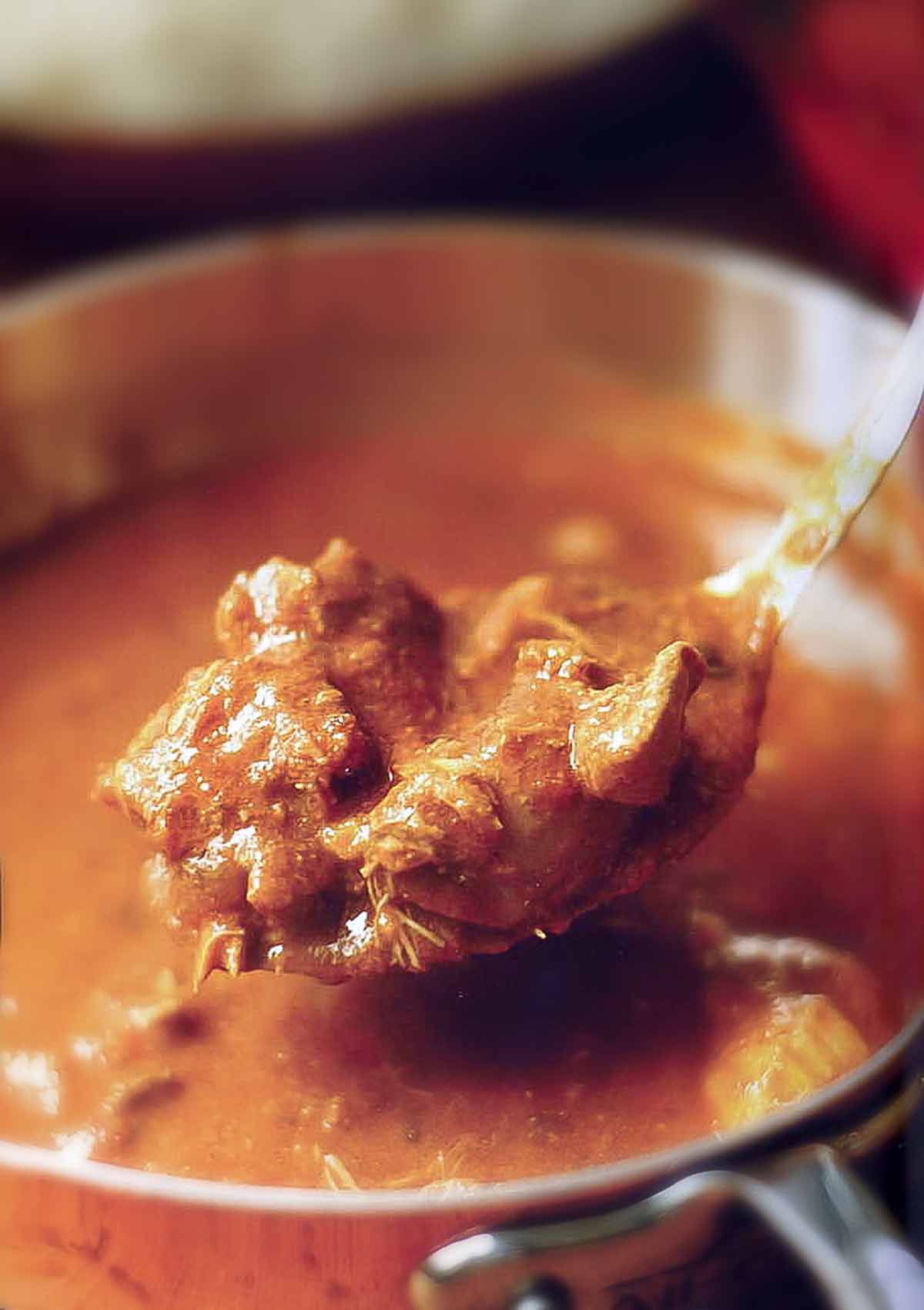 A spoon of a red chicken curry held above copper pot filled with the Indian stew