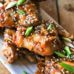 Mahogany wings piled on a white rectangular platter, garnished with scallions and sesame seeds.