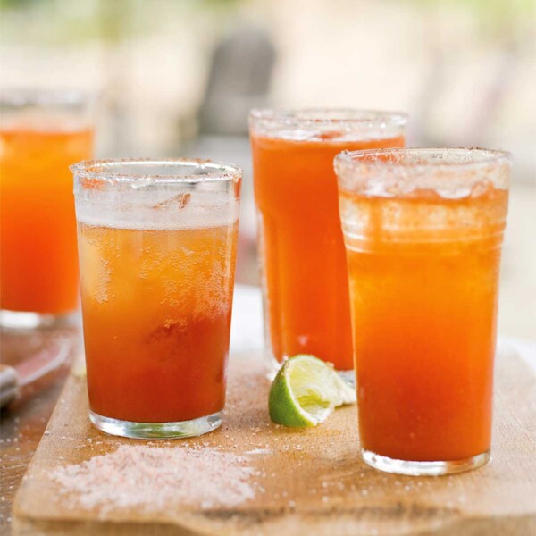 Four glasses of michelada on a wooden board with a lime wedge in between them.