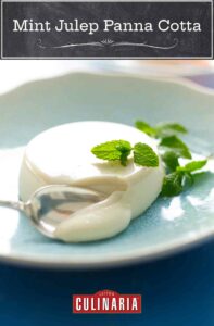 A mint julep panna cotta in a blue bowl, garnished with mint sprigs and a silver spoon scooping it.