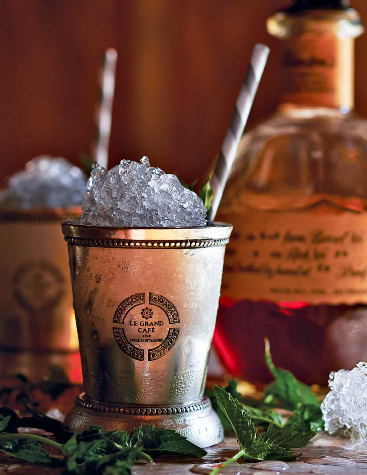 Two copper glasses filled with mint julep with straws sticking out from them and mint leaves scattered around.
