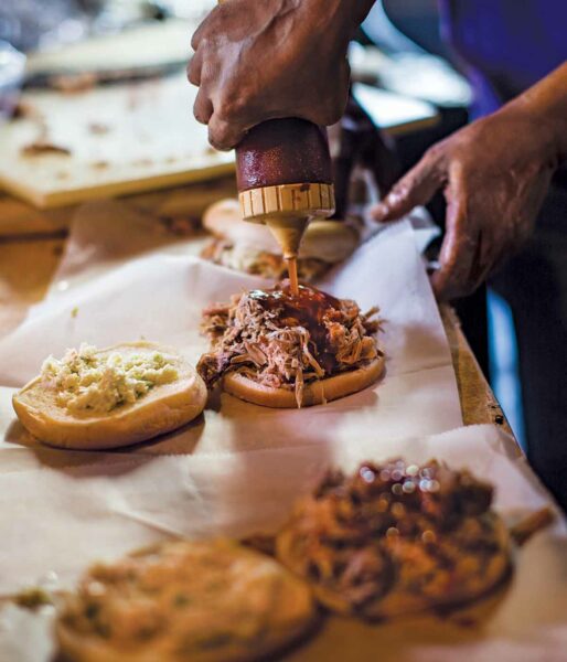 A person squeezing North Carolina barbecue sauce from a plastic bottle onto a sandwich.