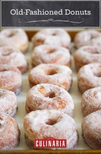 Rows of sugar-coated old-fashioned doughnuts on a rimmed baking sheet.