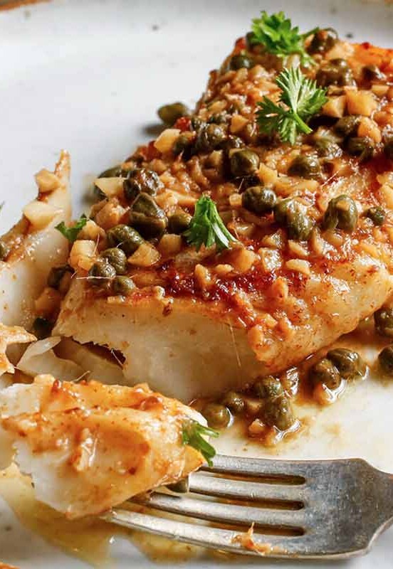 A piece of pan-roasted halibut with caper vinaigrette on a plate with a fork cutting off a piece.