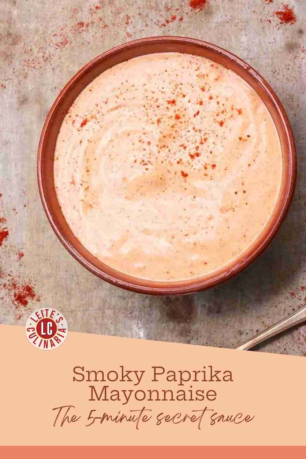 Creamy pale orange paprika mayonnaise in a brown ceramic bowl sprinkled with bright red smoked paprika on a rustic surface.
