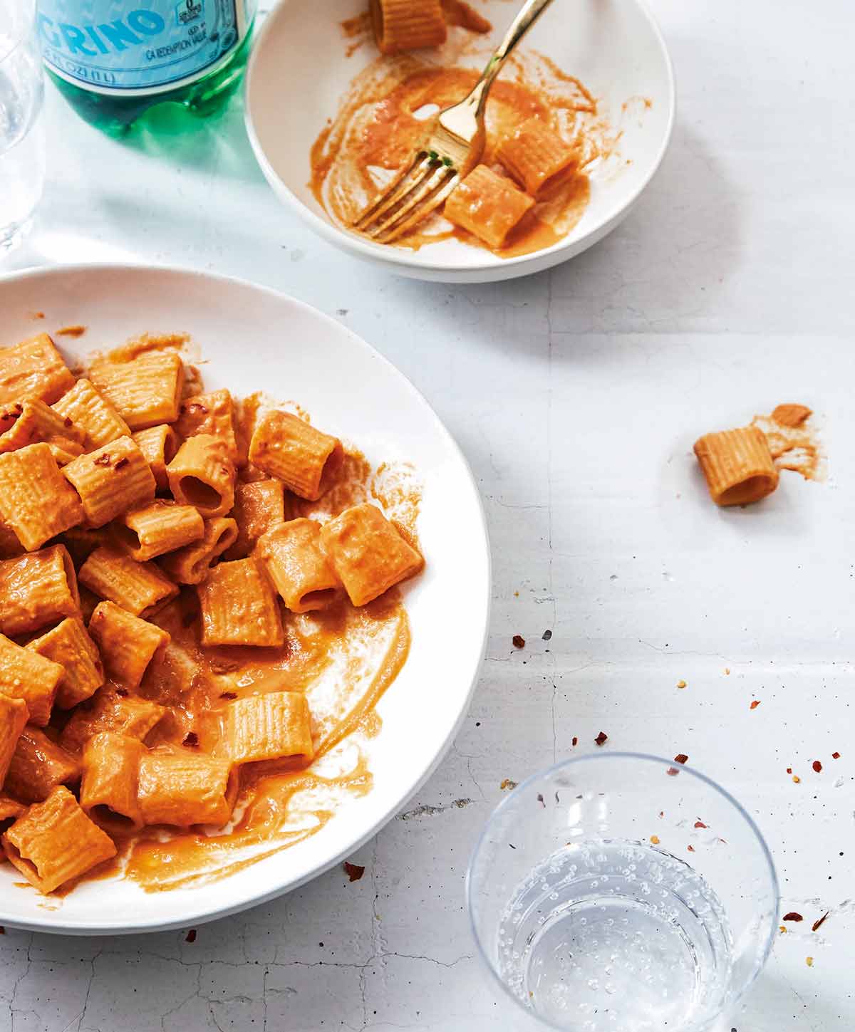 A white serving bowl and smaller white bowl partially filled with rigatoni with vegan vodka sauce and a glass of water on the side.