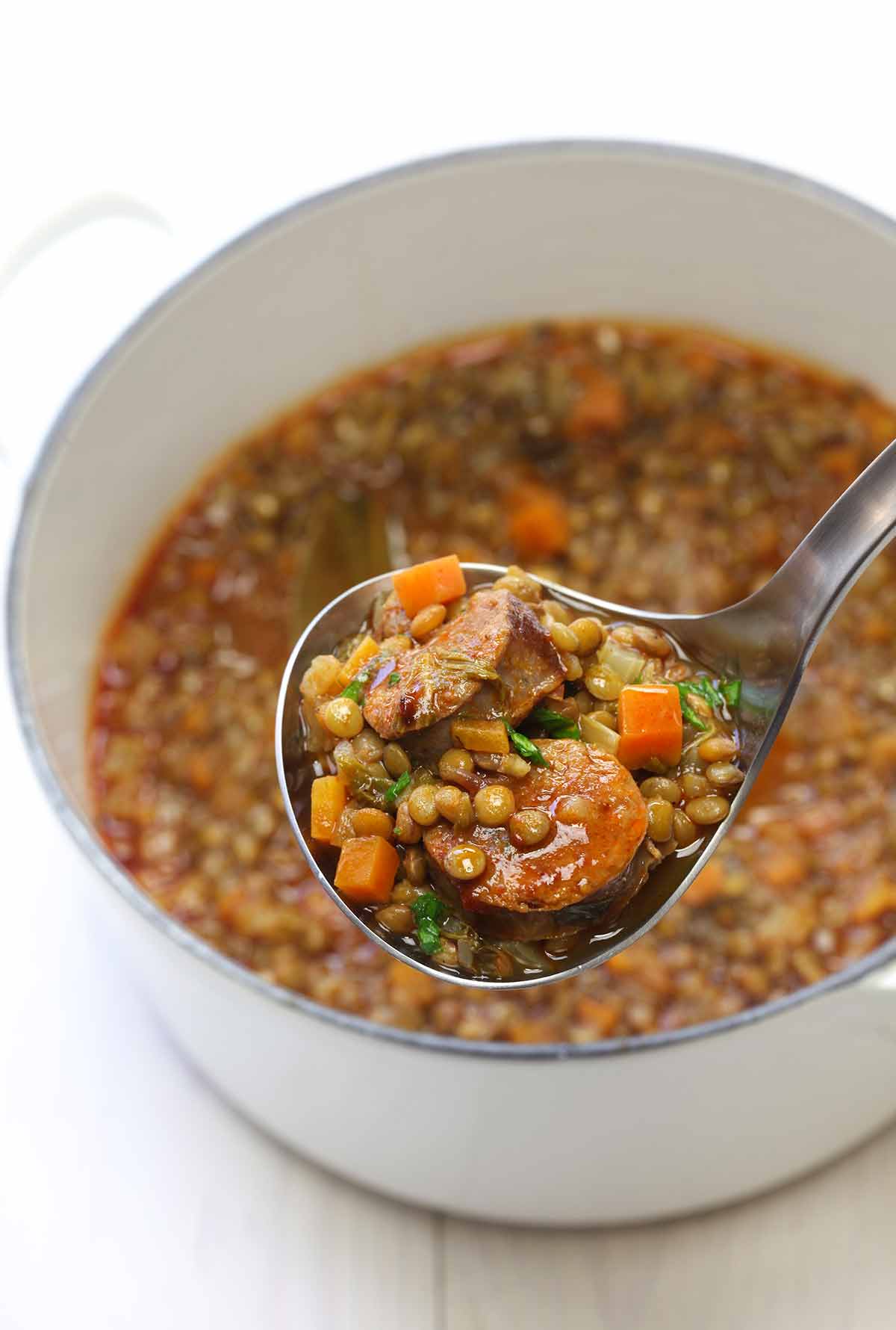 A white pot filled with rustic lentil soup, and a metal ladle scooping out a portion.