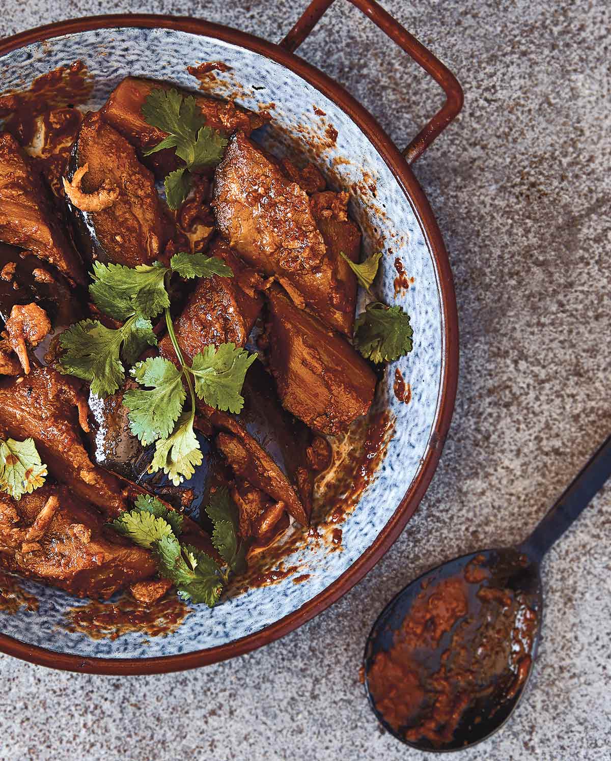 A bowl filled with sambal eggplant, garnished with cilantro, with a black spoon on the side.