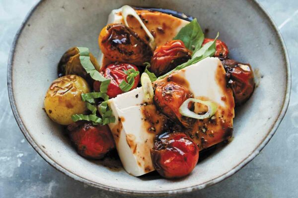 A ceramic bowl filled with silken tofu with soy-sauced tomatoes and scallion and basil leaves to garnish.