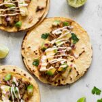 Three assembled slow cooker tacos al pastor with shredded pork, pineapple-avocado salsa, and chipotle mayo, with lime wedges on the side.