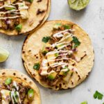 Three assembled slow cooker tacos al pastor with shredded pork, pineapple-avocado salsa, and chipotle mayo, with lime wedges on the side.