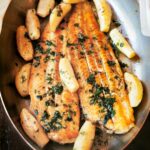 Two filletes of sole à la meunière in an oval metal dish surrounded by lemon wedges and garnished with parsley.